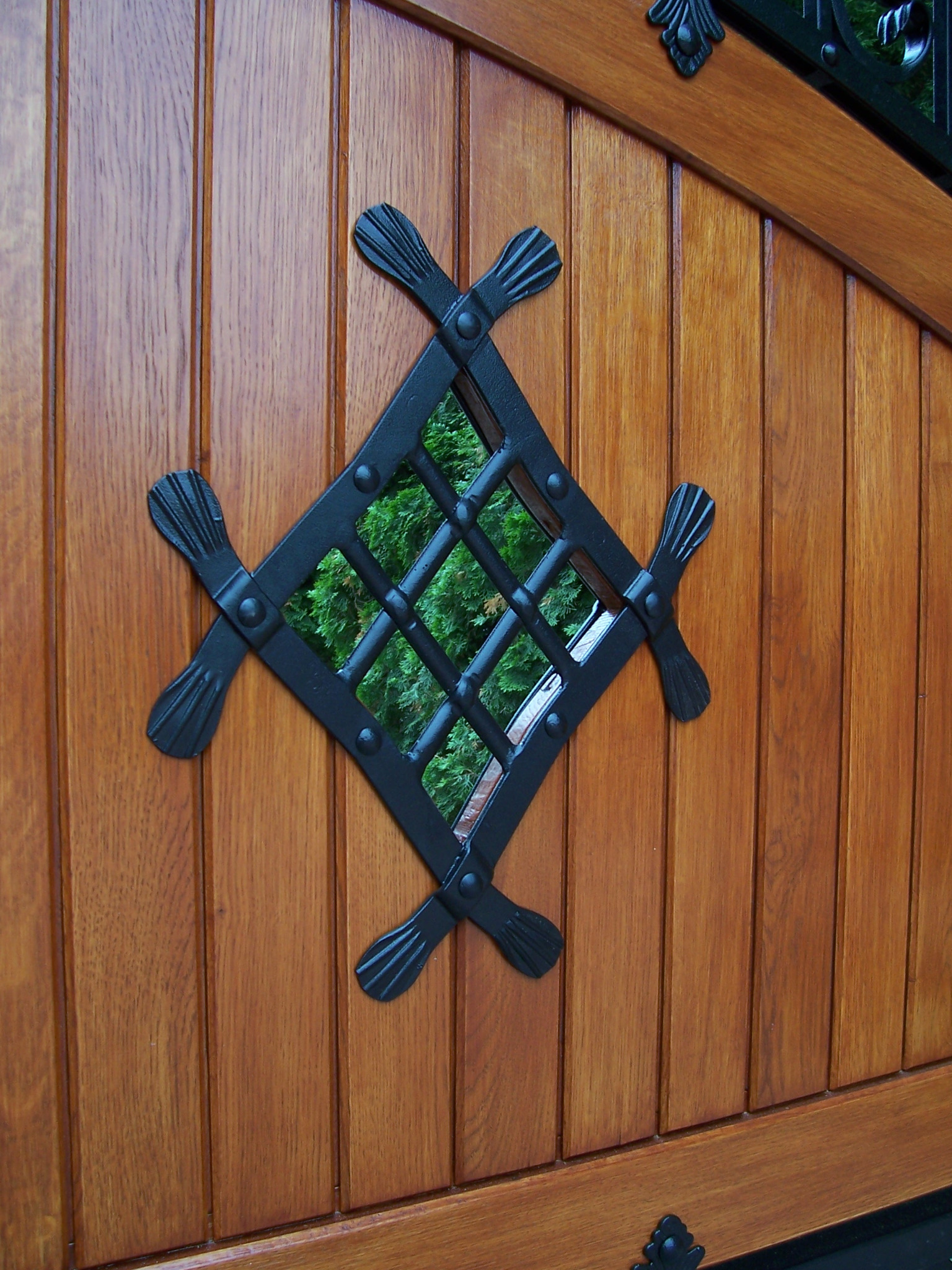 Fragment drewnianej bramy z ozdobnym, kutym okienkiem w kształcie rombu, przez które widać zieleń.