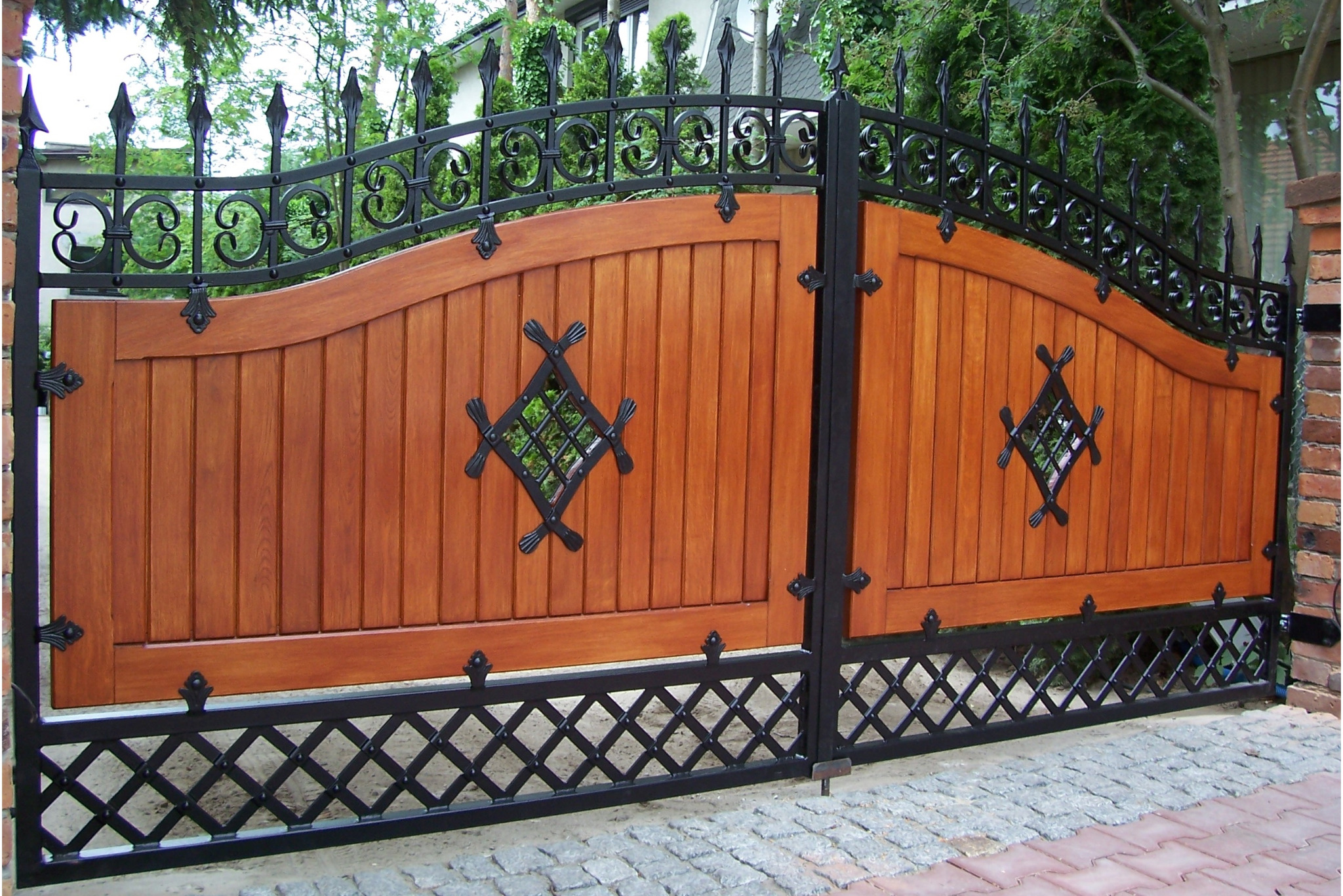 Dwuskrzydłowa brama wjazdowa z drewnianymi panelami w kolorze miodowego dębu, ozdobiona kutymi elementami w kształcie rombów i dekoracyjną, ażurową kratą u dołu, zwieńczona kutym, ozdobnym łukiem...