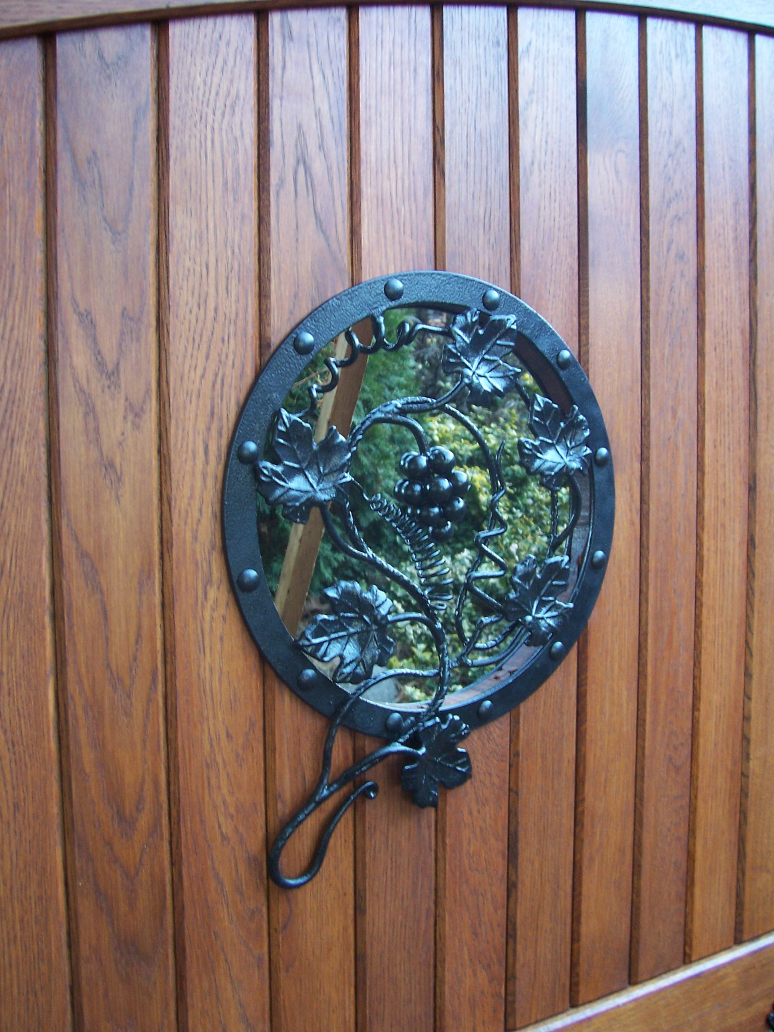 Fragment drewnianej bramy z okrągłym, kutym elementem dekoracyjnym przedstawiającym winogrona i liście, przez który widać zieleń ogrodu.