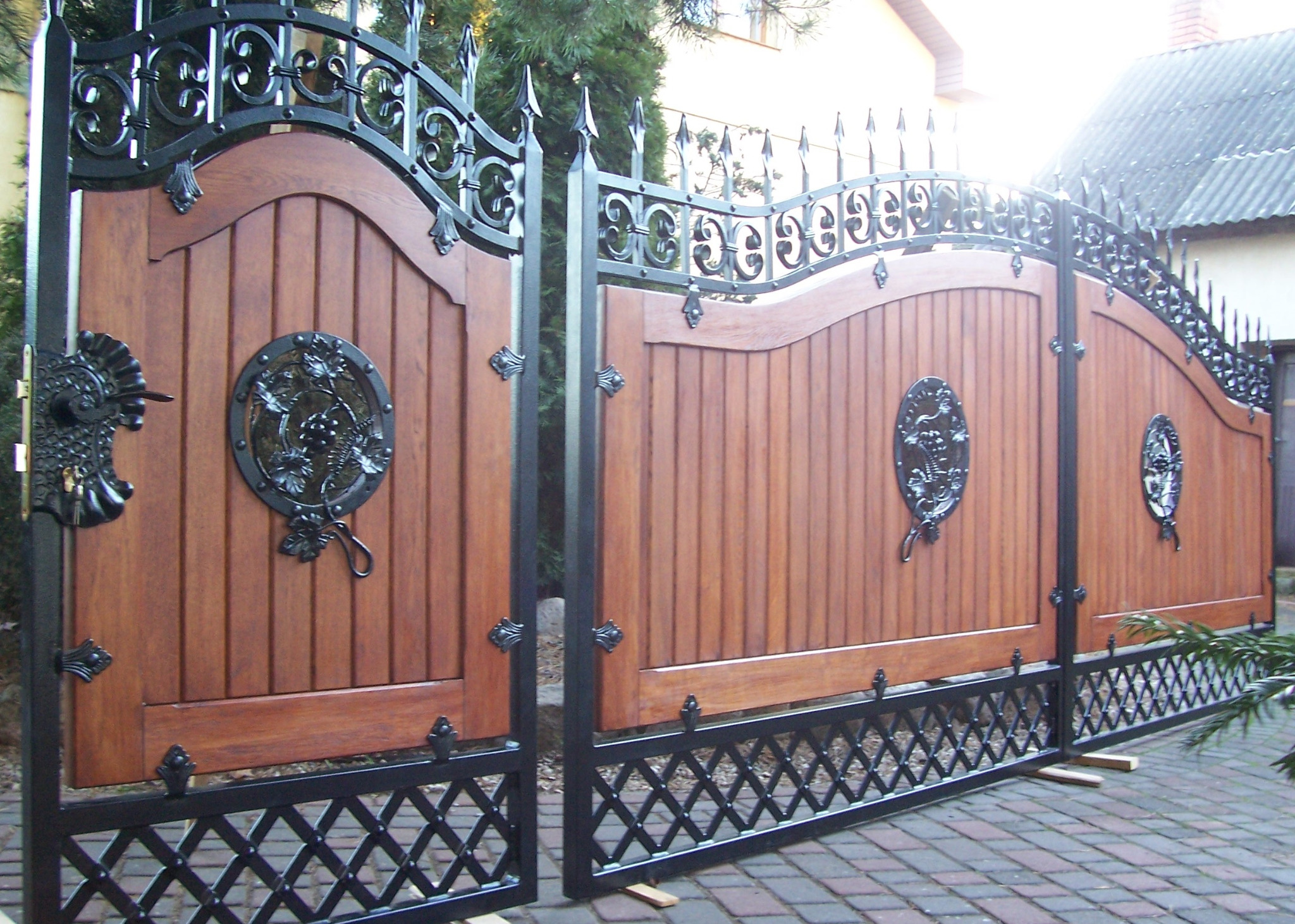 Dwuskrzydłowa brama wjazdowa z drewnianymi panelami ozdobionymi metalowymi rozetami i kutą, ażurową górną częścią, na tle kostki brukowej i zieleni.