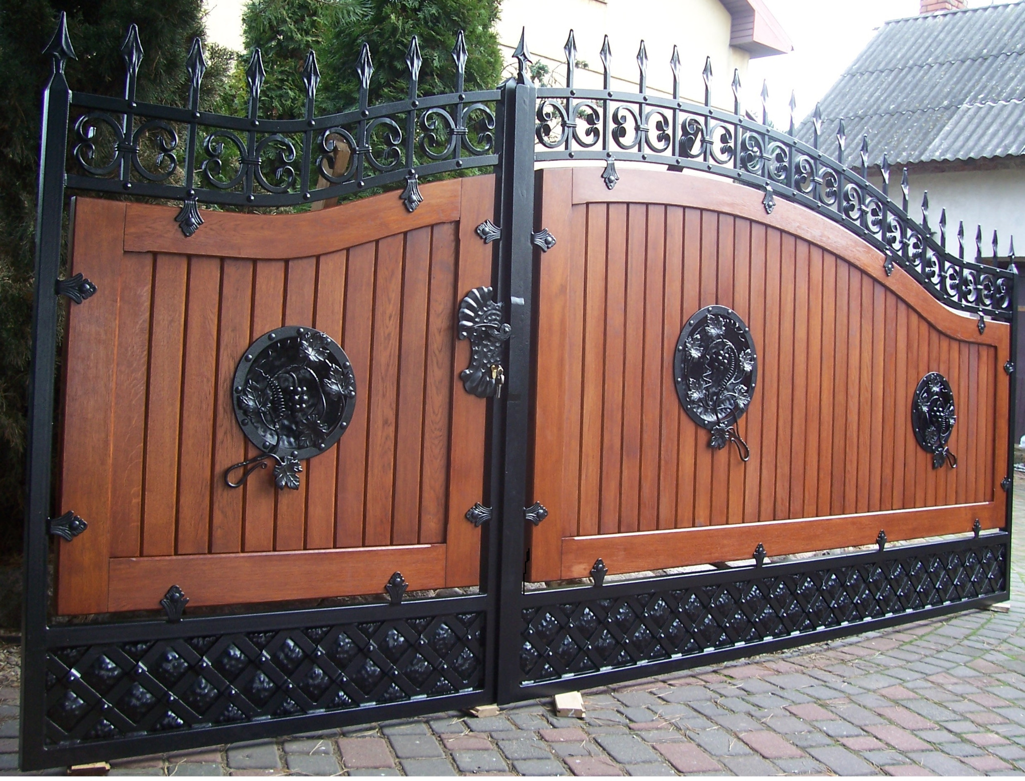 Dwuskrzydłowa brama wjazdowa z drewnianymi panelami w ciepłym odcieniu brązu, ozdobiona kutymi elementami w kolorze czarnym, w tym rozetami z motywem winogron i dekoracyjnym zamknięciem.