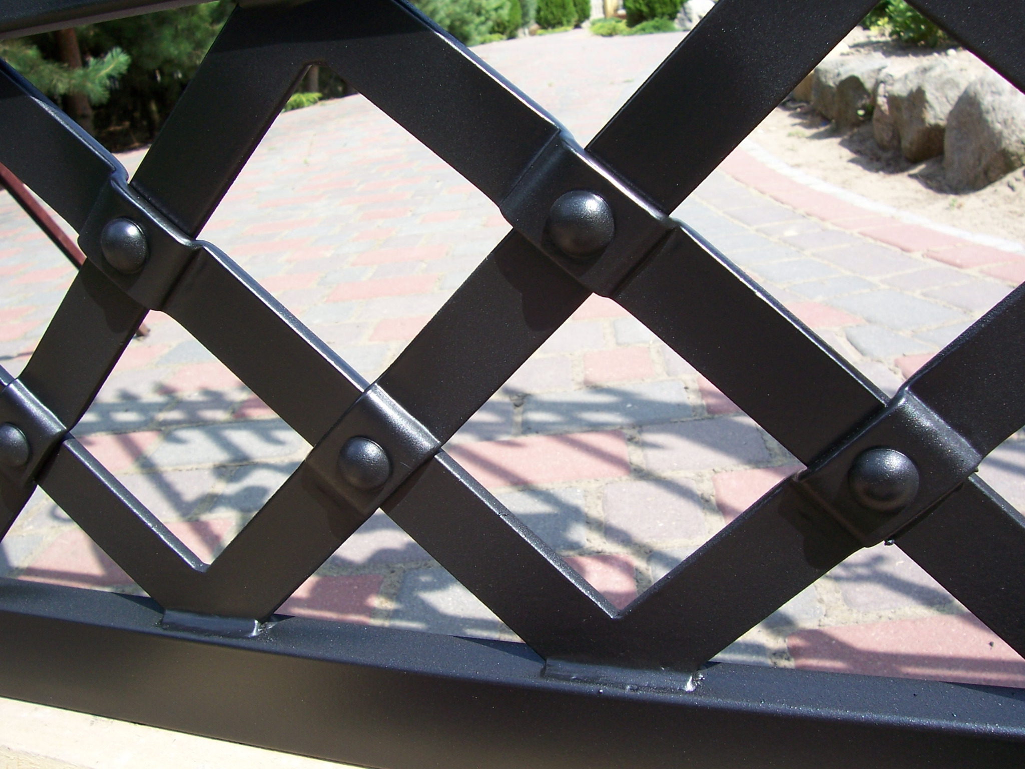 Fragment czarnego, metalowego ogrodzenia o rombowym wzorze, z widocznymi nitami i spoinami, na tle brukowanej ścieżki i zieleni ogrodu.