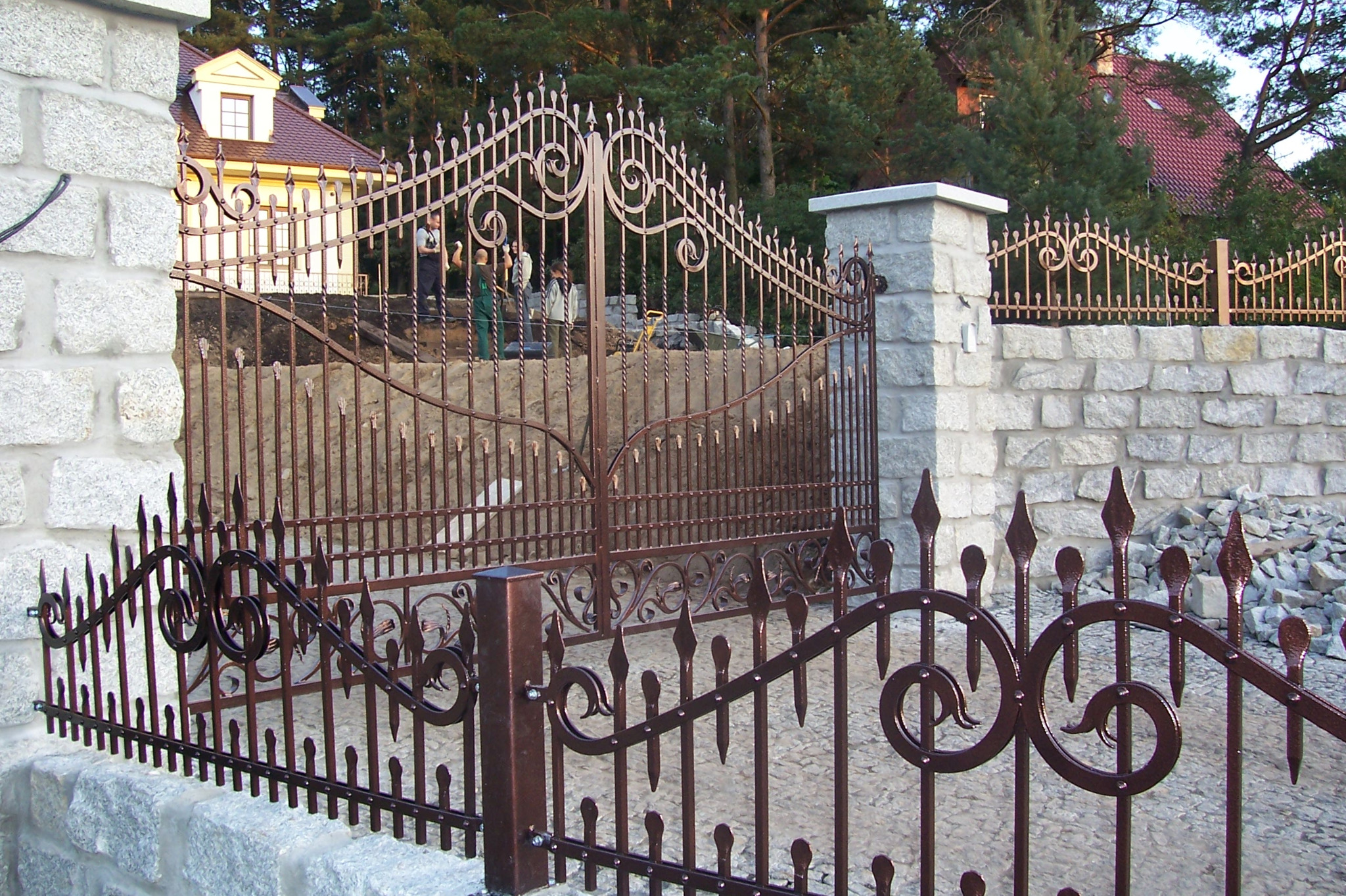 Otwarta, dwuskrzydłowa brama z metaloplastyki w kolorze ciemnej miedzi, z dekoracyjnymi elementami roślinnymi, zamontowana na kamiennych słupkach i niskim murku z kamienia łupanego, w tle widoczni...