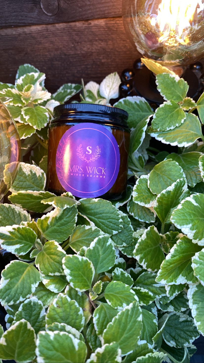Sojowa świeca zapachowa Mrs. Wick w brązowym słoiczku z fioletową etykietą, otoczona zielonymi liśćmi z białymi brzegami, w tle żarząca się żarówka dekoracyjna.