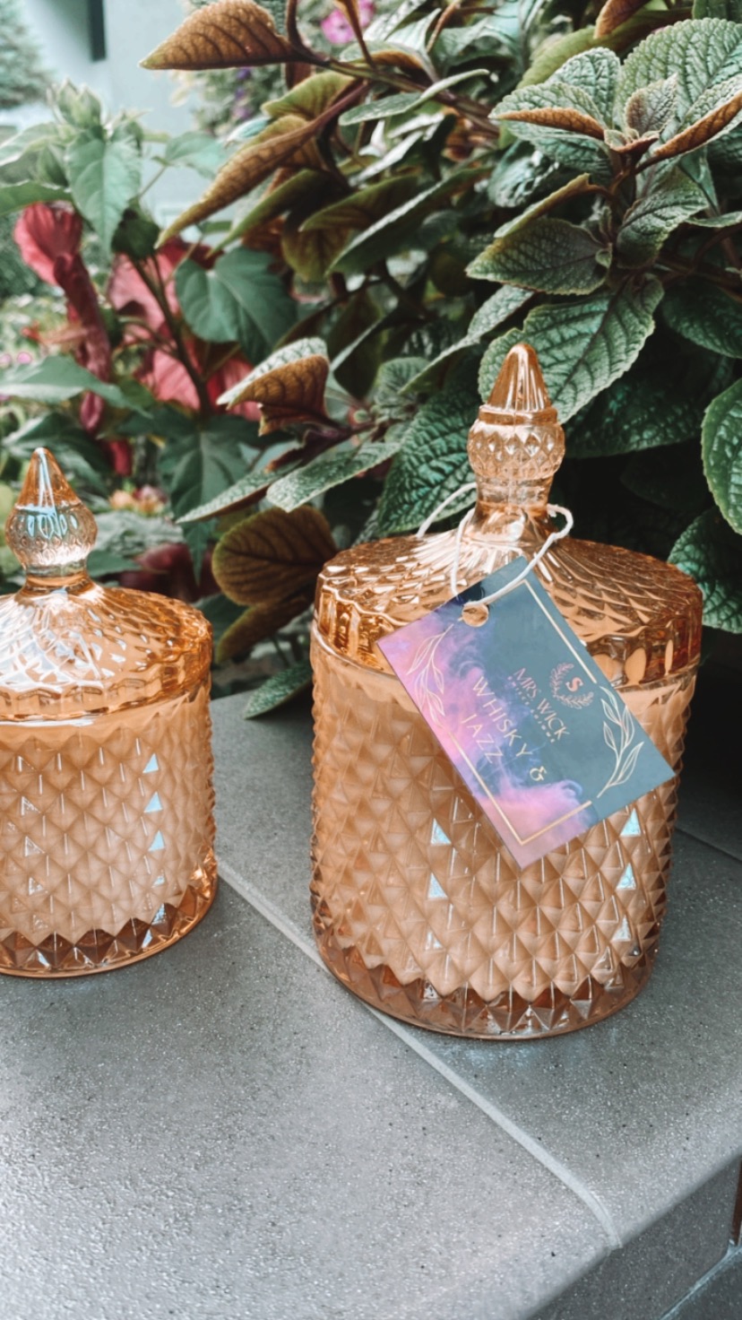 Dwa dekoracyjne, złote słoiki z geometrycznym wzorem i wieczkami w kształcie stożka, z zawieszką z napisem 'Whisky & Jazz' na tle roślinności.