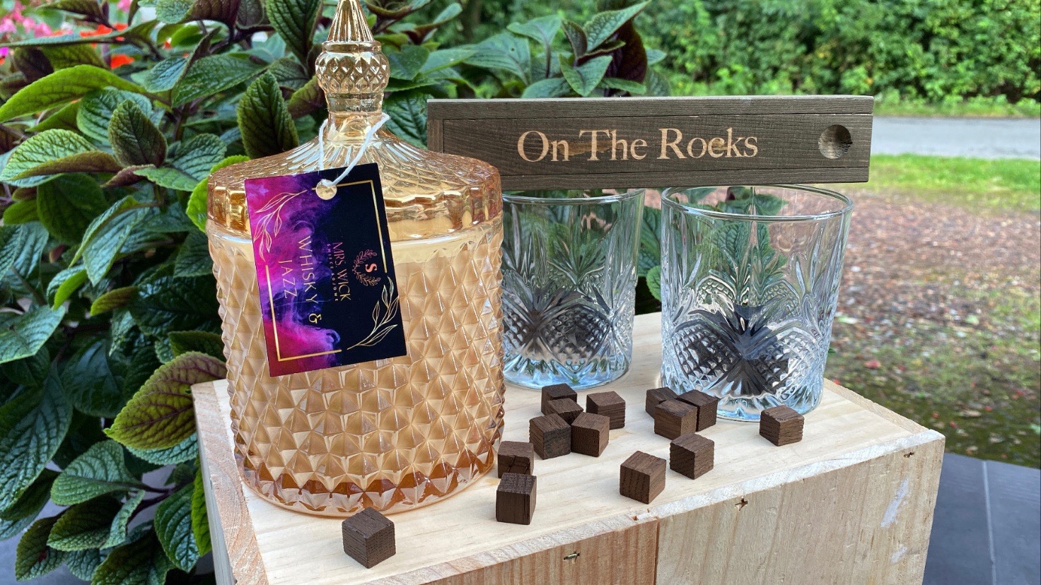 Zestaw prezentowy: zdobiona szklana świeca zapachowa z etykietą 'Whisky & Jazz', dwa szklane kieliszki z wzorem i drewniane kostki na drewnianej skrzynce, z napisem 'On The Rocks' w tle.