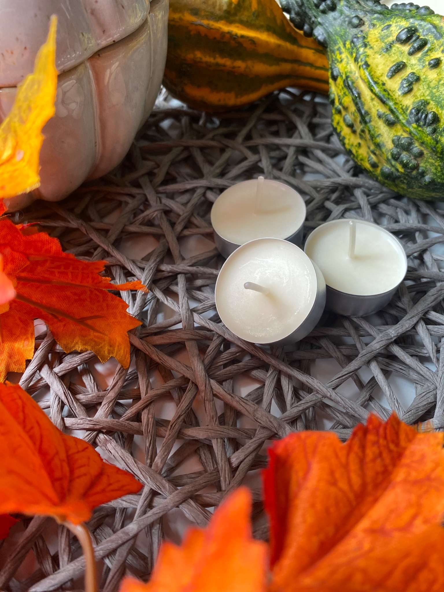 Jesienna kompozycja z trzema białymi świeczkami typu tealight, dekoracyjnymi dyniami i pomarańczowymi liśćmi na plecionym, szarym podkładce.