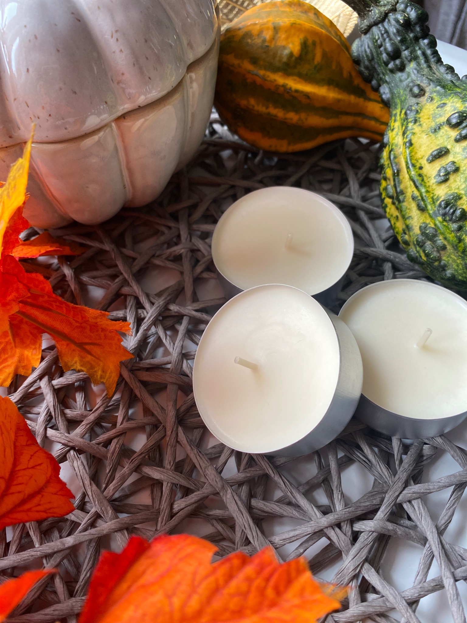 Jesienna kompozycja z dekoracyjną białą dynią, ozdobnymi tykwami, trzema świeczkami typu tealight i pomarańczowymi liśćmi na plecionym podkładce.