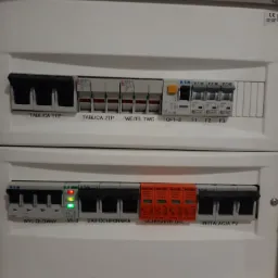 Szczegółowe ujęcie dwupoziomowej rozdzielnicy elektrycznej z widocznymi bezpiecznikami, wyłącznikiem głównym i zabezpieczeniami przepięciowymi, w tym pomarańczowymi ogranicznikami B+C, z opisami...