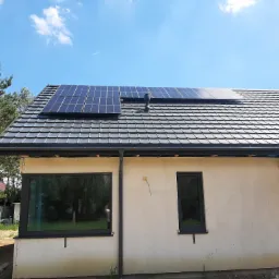 Panele fotowoltaiczne zamontowane na dachu domu jednorodzinnego z szarą dach&oacute;wką, widok z dołu na tle błękitnego nieba, instalacja w trakcie wykańczania elewacji.