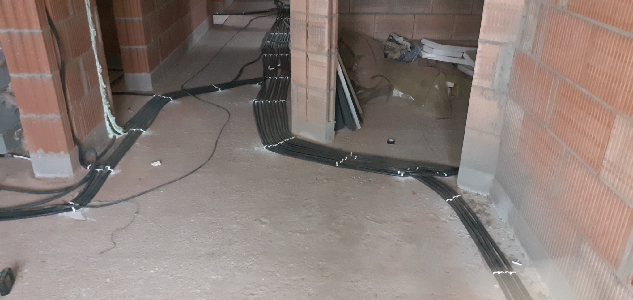 Wiązka przewodów elektrycznych ułożona na podłodze w surowym stanie, mocowana do ściany z cegły, widoczne elementy konstrukcyjne budynku.