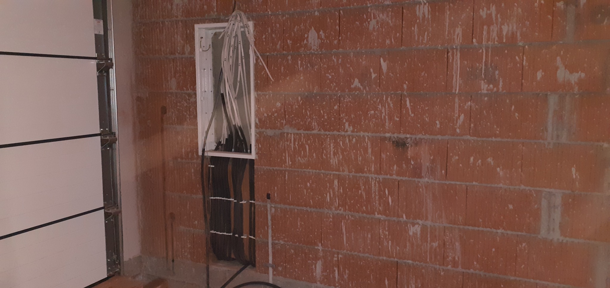 Surowa ściana z czerwonej cegły z otwartą białą skrzynką elektryczną, z której wystają liczne kable w kolorach białym i czarnym, częściowo ukryte w ścianie. Obok widoczny fragment białej bramy...