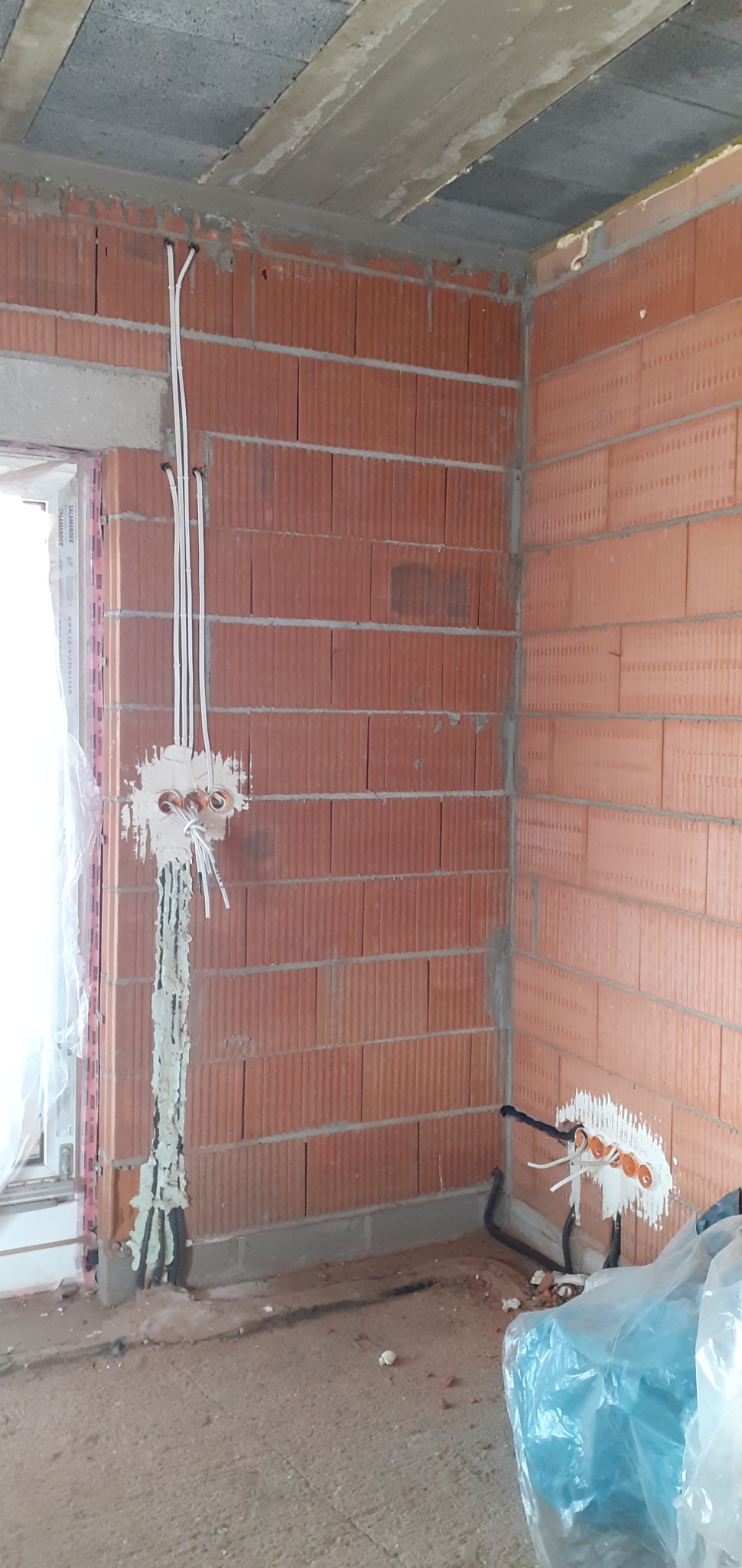 Wewnętrzny róg budynku w trakcie budowy z widocznymi przewodami elektrycznymi wychodzącymi ze ściany, zabezpieczonymi pianką montażową i gipsem.