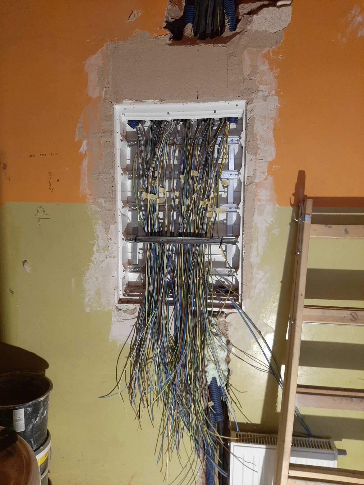 Rozdzielnica elektryczna w trakcie montażu w ścianie, widoczne liczne przewody, drabina oparta o ścianę, surowe wykończenie wnętrza.