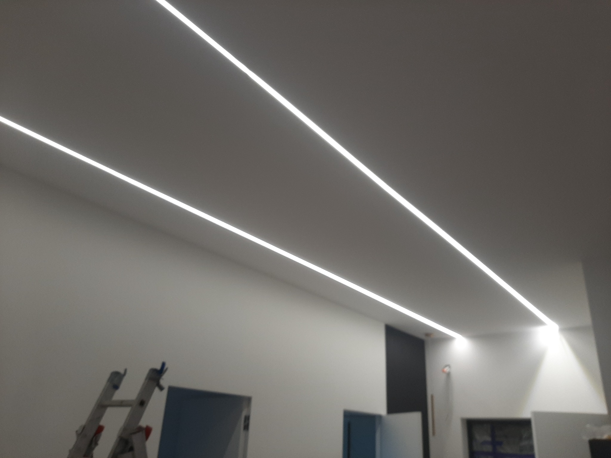 Dwie równoległe linie oświetlenia LED zamontowane w suficie podwieszanym, widoczna drabina aluminiowa w tle.