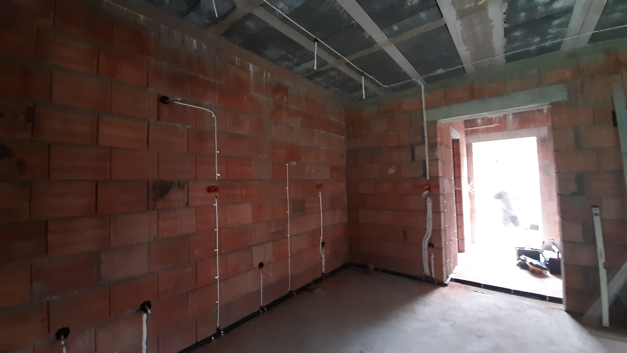 Wnętrze w stanie surowym z widoczną instalacją elektryczną: przewody poprowadzone wzdłuż ceglanej ściany, puszki elektryczne i peszle przy podłodze.