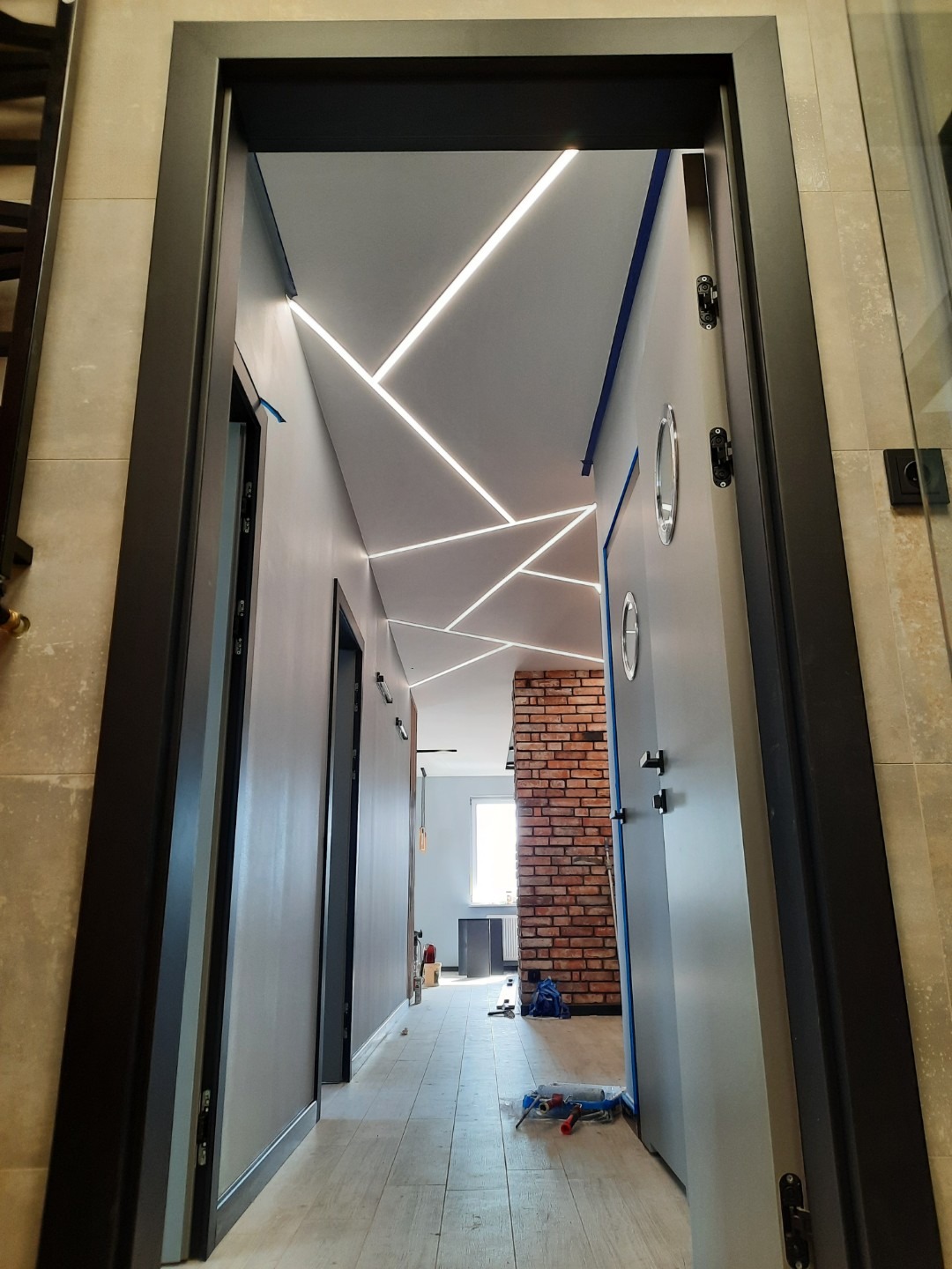 Nowoczesny korytarz w Ślądkowicach z geometrycznym oświetleniem LED na suficie, widok zza futryny drzwi, narzędzia malarskie na podłodze.