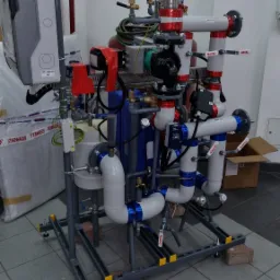 Złożona instalacja hydrauliczna z licznymi rurami, zaworami i pompami, zamontowana na stalowej ramie, ustawiona na cegłach na podłodze z płytek. Widoczne elementy elektryczne i oznaczenia...