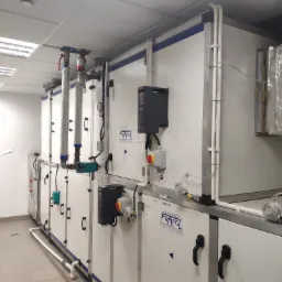 Długa instalacja wentylacyjna FRAPOL w pomieszczeniu technicznym, widoczne rury, zawory, czarne uchwyty i elementy sterujące, oświetlenie sufitowe.