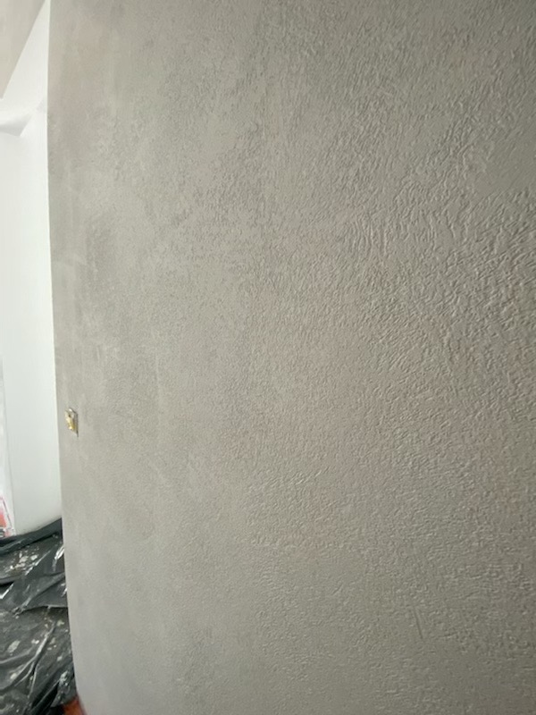 Ściana z szarą, dekoracyjną strukturą, widoczny fragment narożnika i folia ochronna na podłodze.