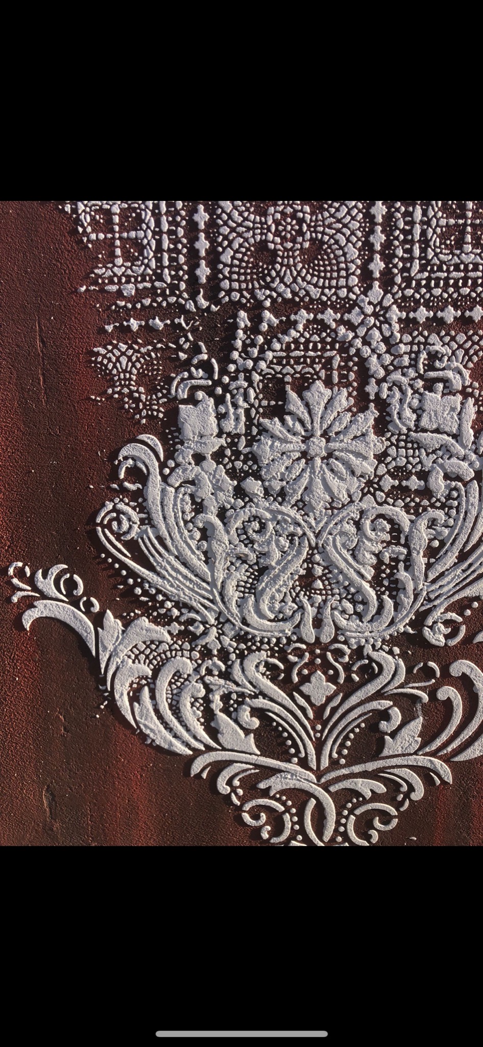 Dekoracyjny tynk strukturalny w kolorze ceglastym z wypukłym, białym ornamentem roślinnym i geometrycznym, oświetlony z góry.