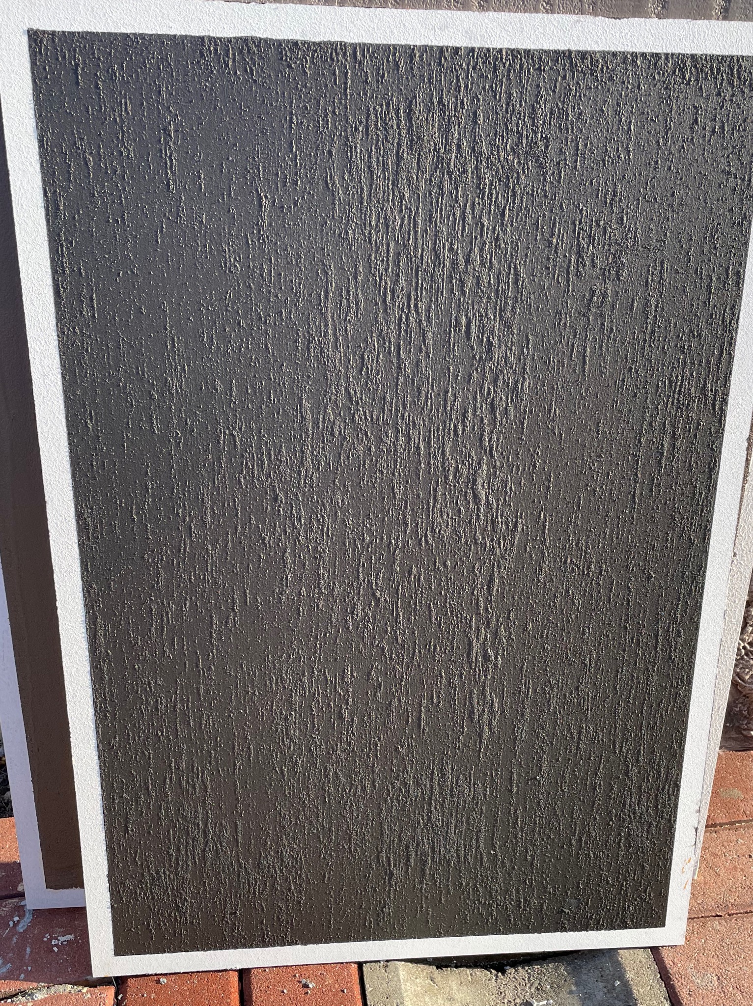 Próbka ciemnoszarej, strukturalnej wyprawy tynkarskiej z wyraźnym pionowym wzorem, oprawiona w białą ramę, ułożona na ceglanej powierzchni.