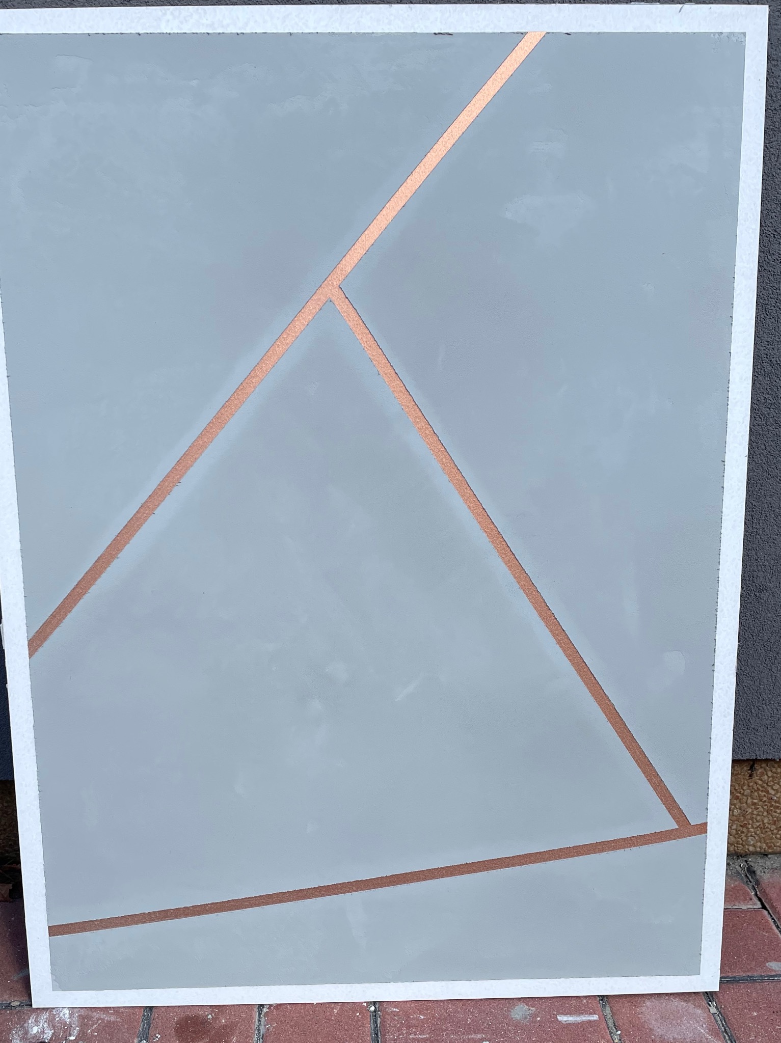 Szara ściana z dekoracyjnymi, miedzianymi paskami tworzącymi geometryczny wzór, wykończona białą ramą.