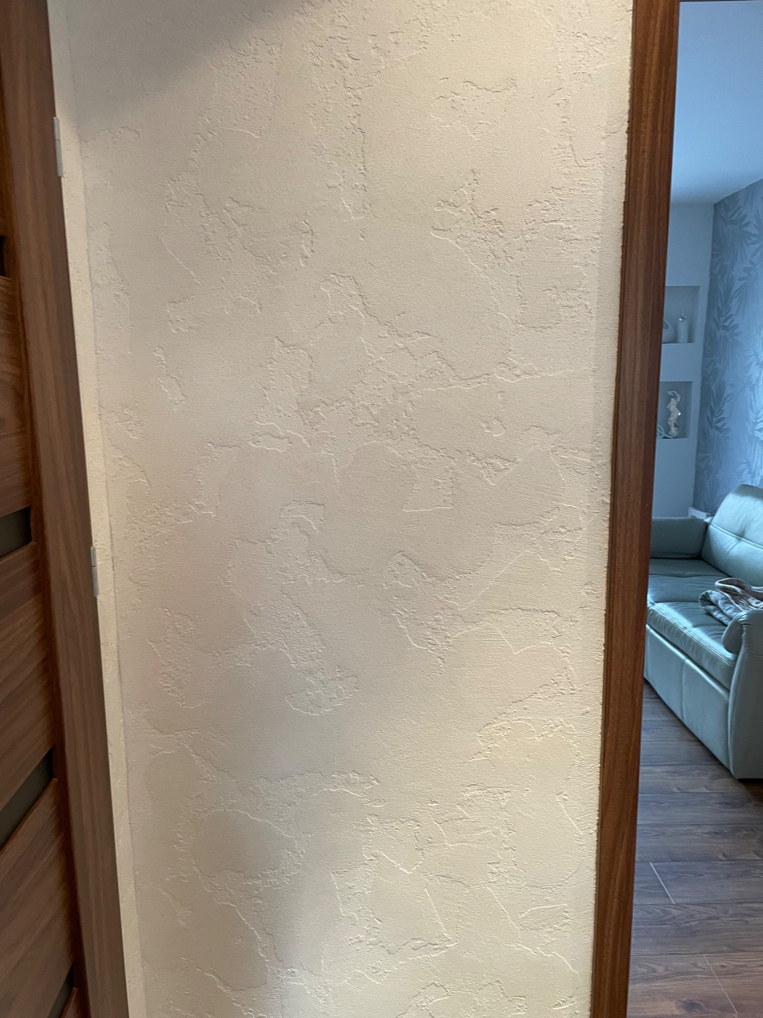 Ściana z dekoracyjną, jasną strukturą w korytarzu, widoczna przez framugę drzwi z drewnianym wykończeniem. W tle fragment salonu z jasną sofą.