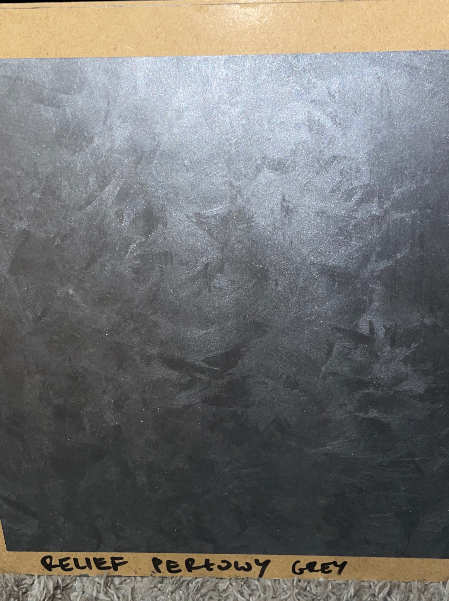 Próbka szarej, perłowej farby strukturalnej o reliefowej fakturze, umieszczona na kartonowej podkładce z odręcznym napisem 'Relief Perłowy Grey'.