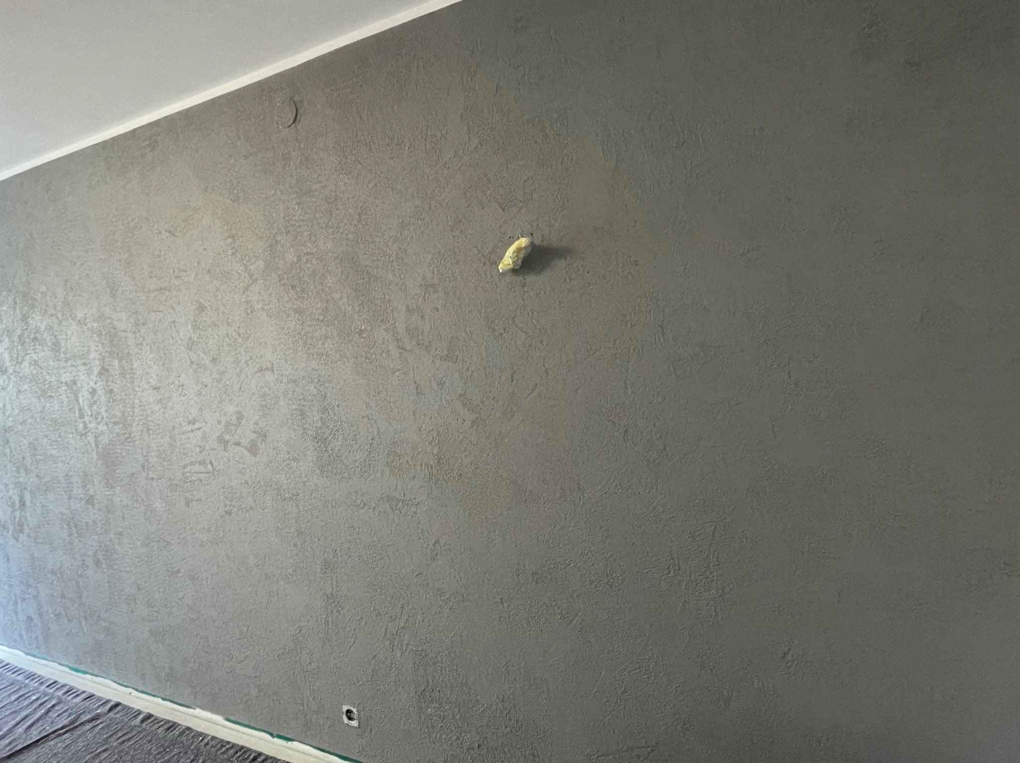 Szara ściana z fakturą po nałożeniu tynku dekoracyjnego, zabezpieczona gniazdkiem elektrycznym i fragmentem sufitu z listwą, widoczne niedoskonałości i zakryta podłoga folią ochronną.