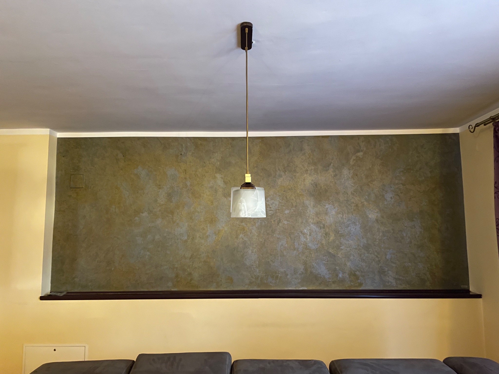 Dekoracyjna ściana z efektem stiuku w odcieniach brązu i szarości, oddzielona od żółtej ściany ciemnobrązową listwą, oświetlona wiszącą lampą z białym kloszem.