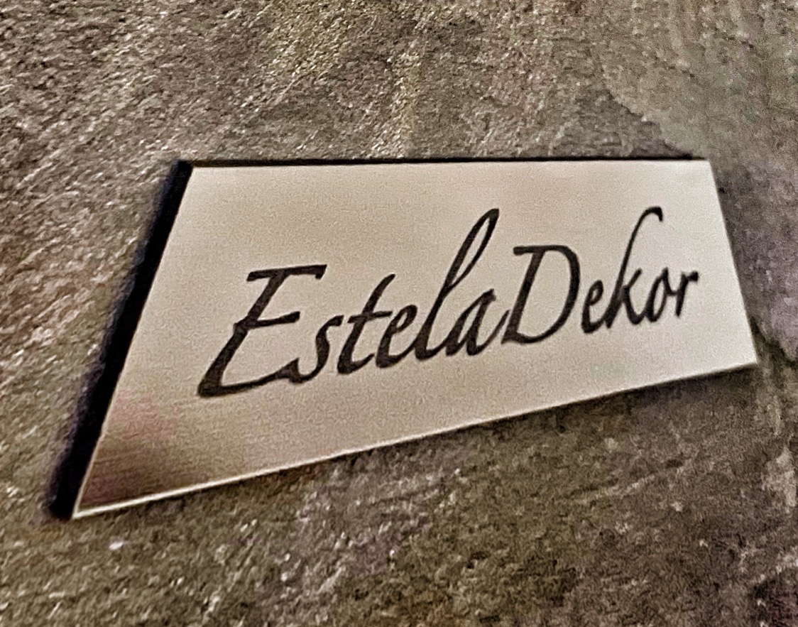 Metalowa tabliczka z napisem 'Estela Dekor' na szarej, teksturowanej ścianie.
