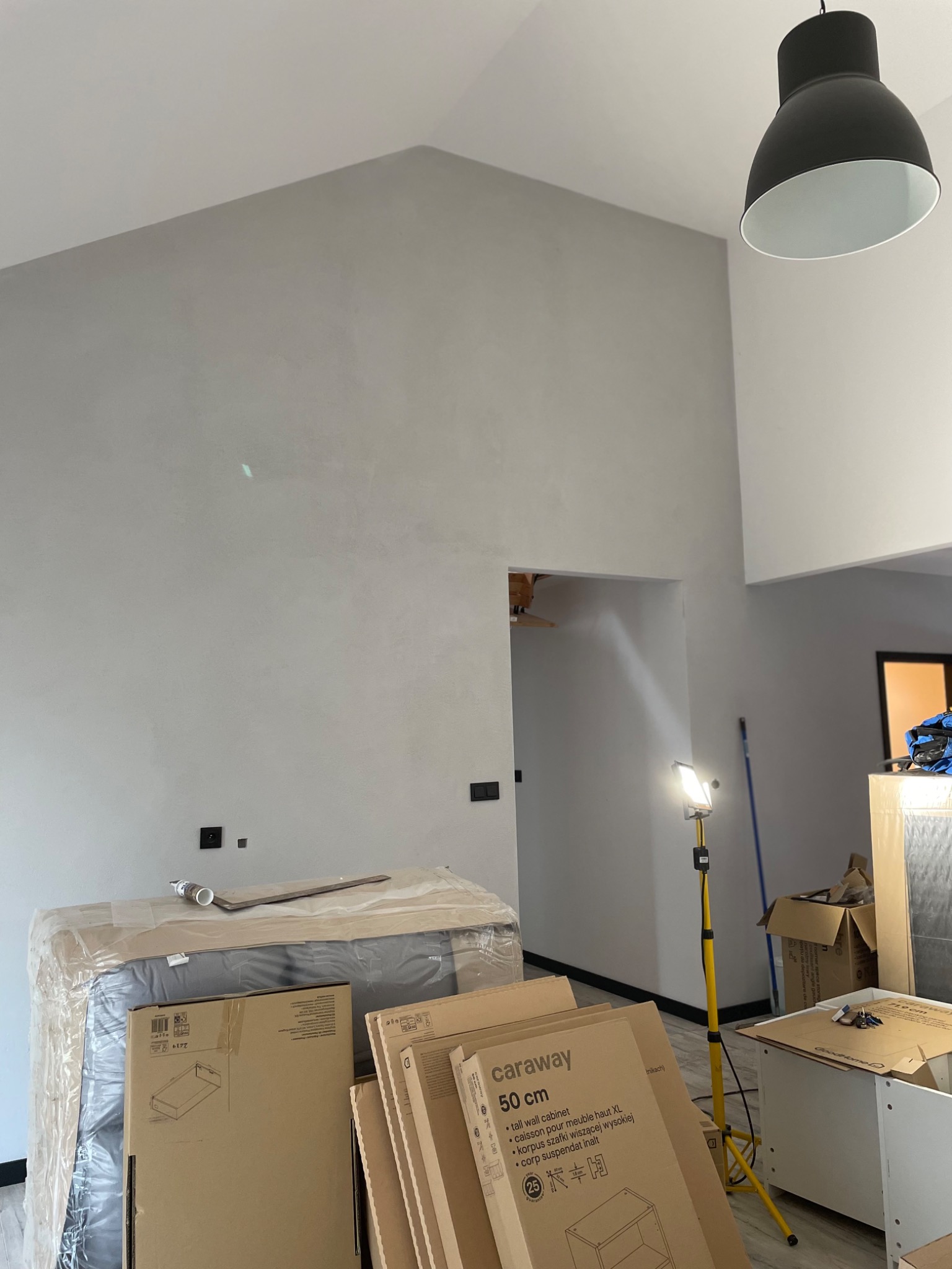 Wnętrze w trakcie wykończenia, ściana pokryta szarą gładzią, widoczne puszki elektryczne, lampa sufitowa i kartony meblowe Caraway 50cm. Na podłodze lampa budowlana na statywie.
