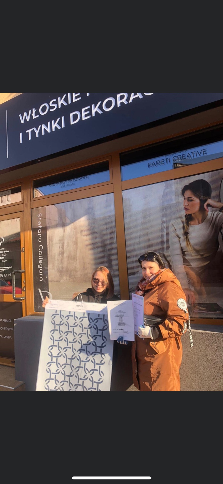 Dwie kobiety przed witryną firmy z włoskimi dekoracjami i tynkami, jedna trzyma szablon dekoracyjny z geometrycznym wzorem, a druga certyfikat.