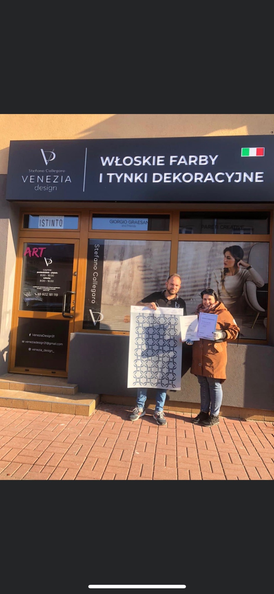 Dwie osoby stojące przed wejściem do firmy 'Venezia Design' z szyldem 'Włoskie farby i tynki dekoracyjne', prezentują szablon z geometrycznym wzorem i certyfikat.