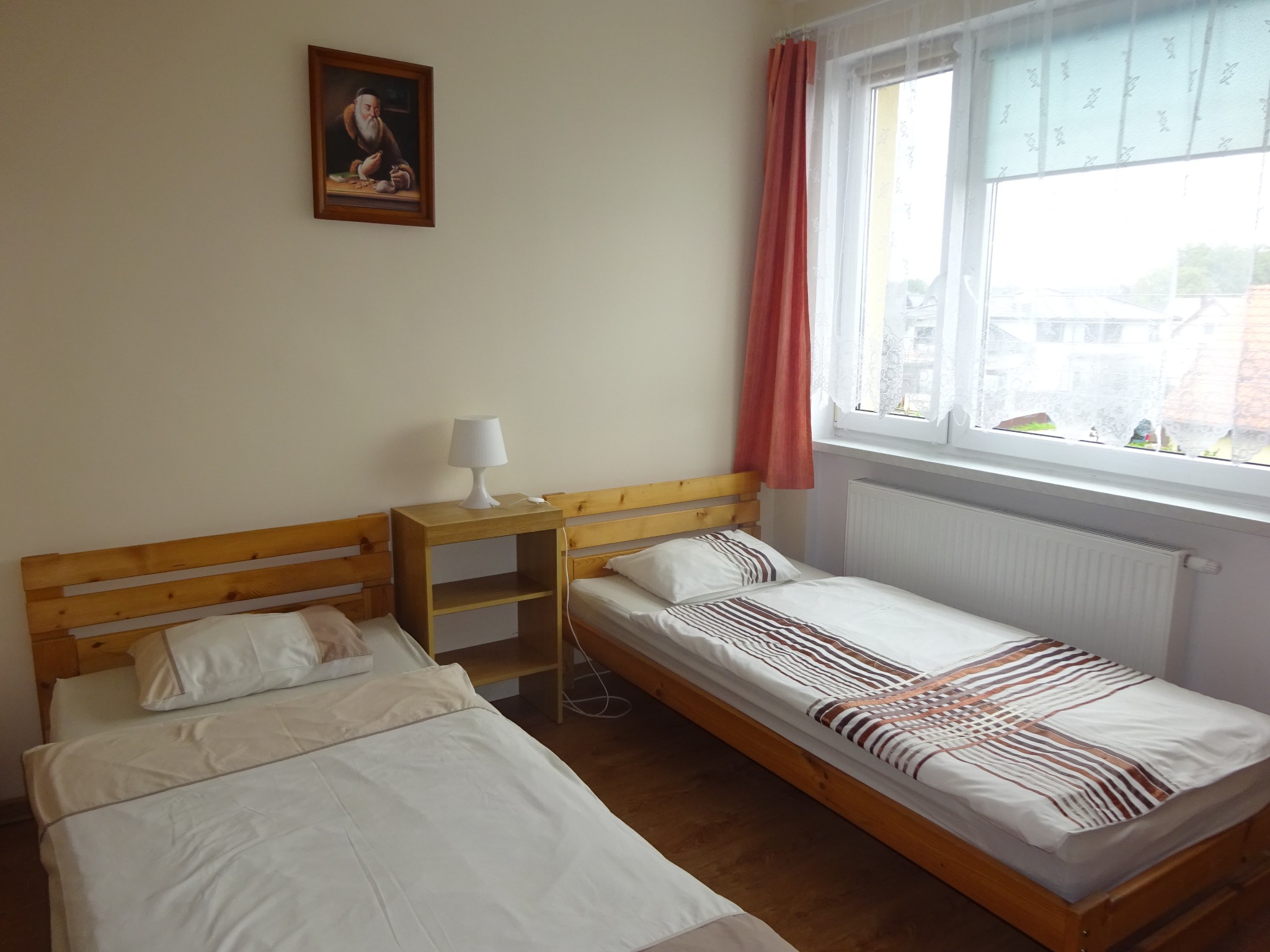 Pokój z dwoma pojedynczymi łóżkami z drewnianymi ramami, mała szafka nocna z lampką, obraz na ścianie i okno z zasłonami.