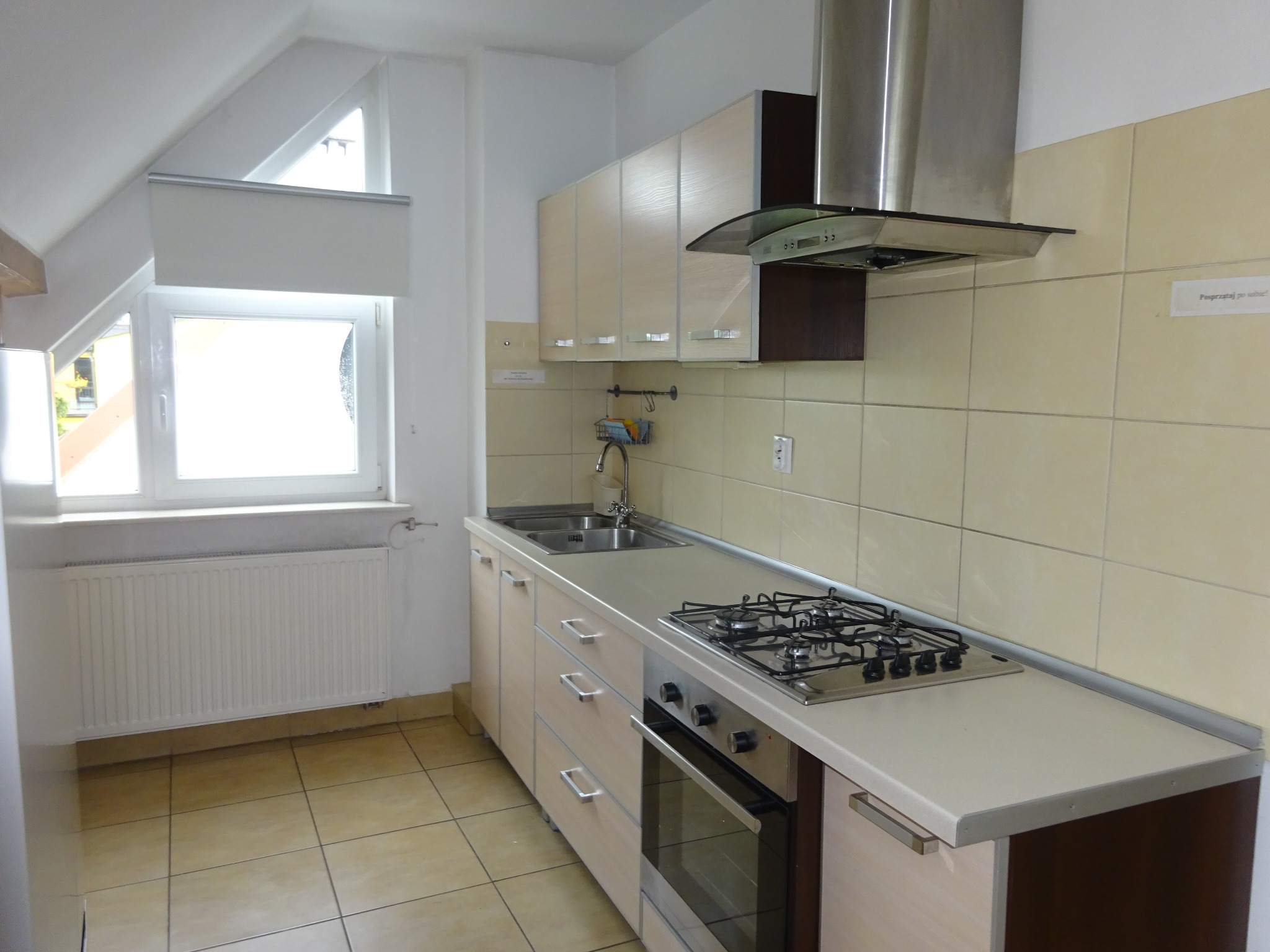 Jasna kuchnia w wynajmowanym mieszkaniu z płytą gazową, zlewozmywakiem i okapem, widok z niskiego kąta, widoczne okno dachowe.