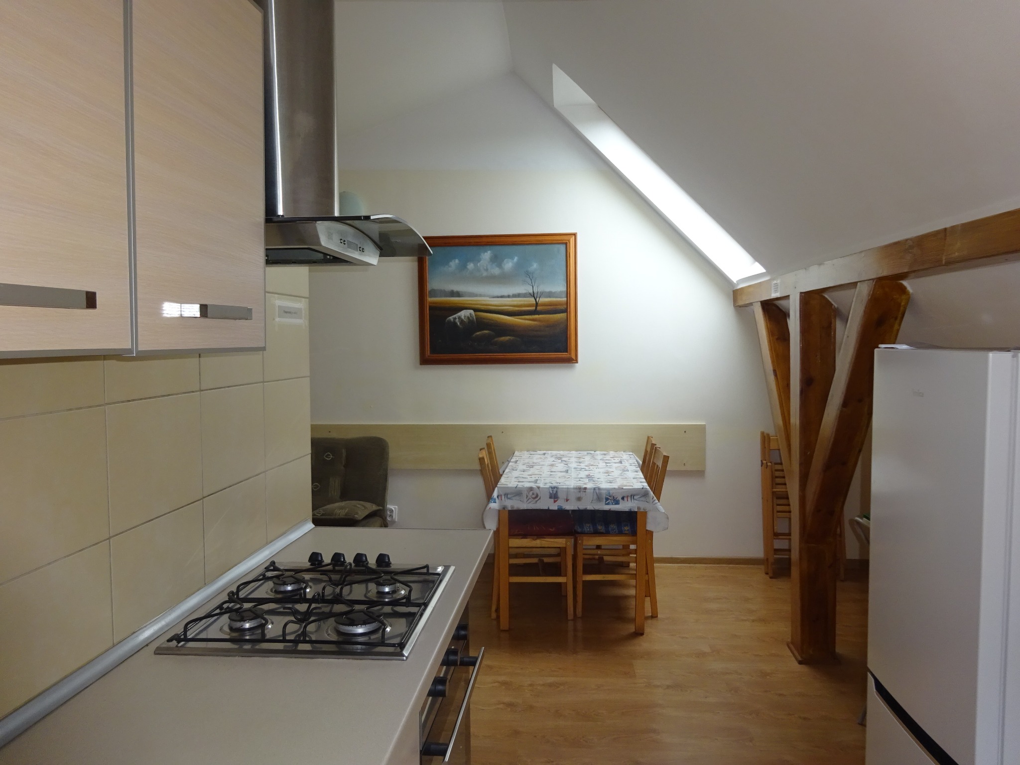 Wnętrze kuchni w wynajmowanym apartamencie na poddaszu z oknem dachowym, widok na stół z krzesłami, płytę grzewczą i obraz na ścianie.