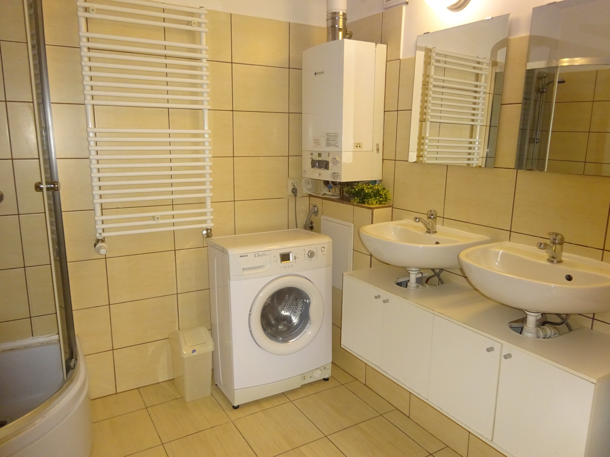 Jasna łazienka z dwiema umywalkami, pralką i grzejnikiem drabinkowym, wykończona beżowymi płytkami.