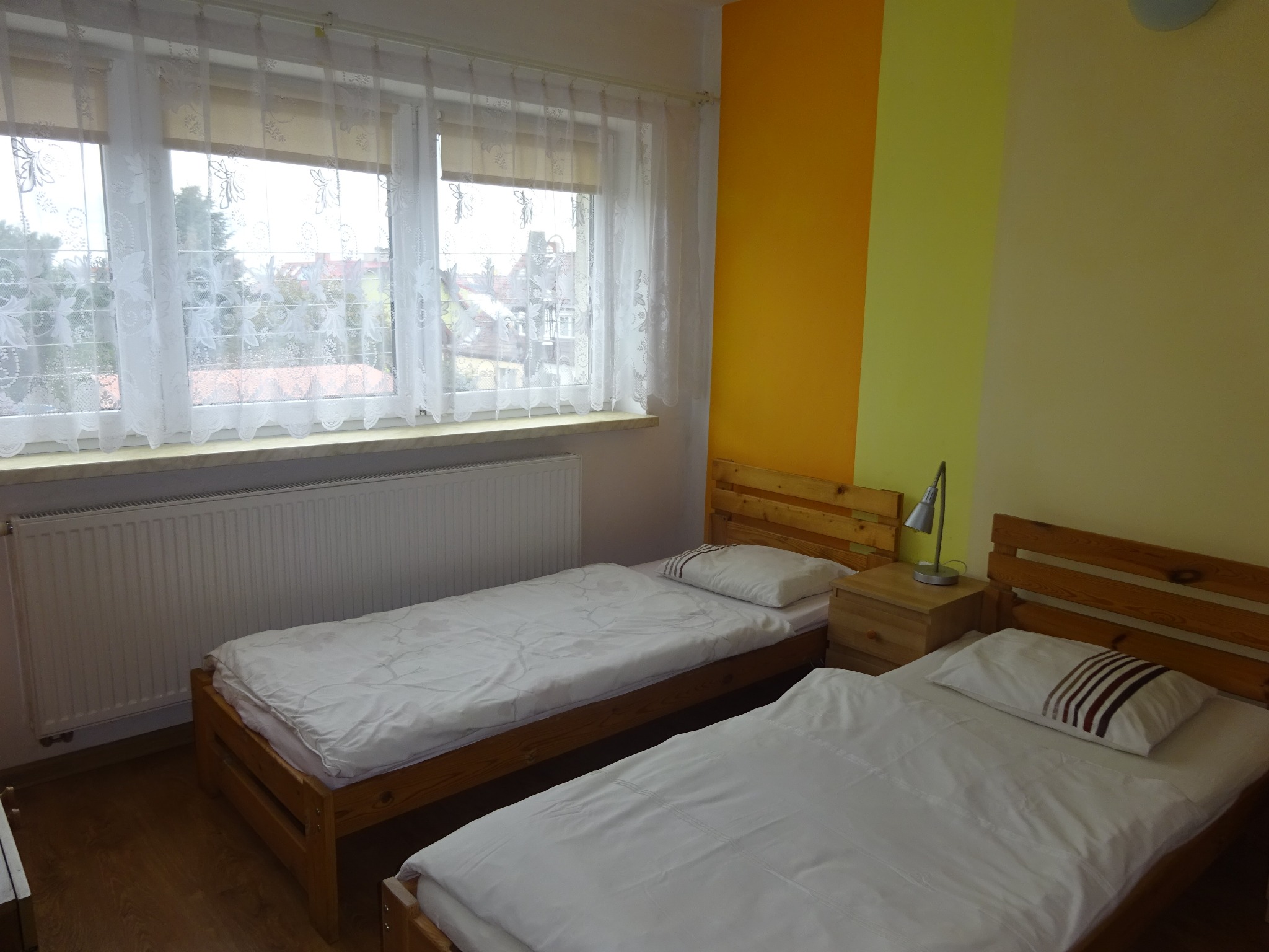 Pokój z dwoma łóżkami pojedynczymi, drewnianymi ramami, białymi pościelami i poduszkami, oświetlony lampką nocną na szafce, z widokiem na zewnątrz przez okno z firankami.