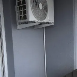 Biała jednostka klimatyzacji Hyundai zamontowana na szarej ścianie, widoczny fragment drabiny i rury instalacyjne.