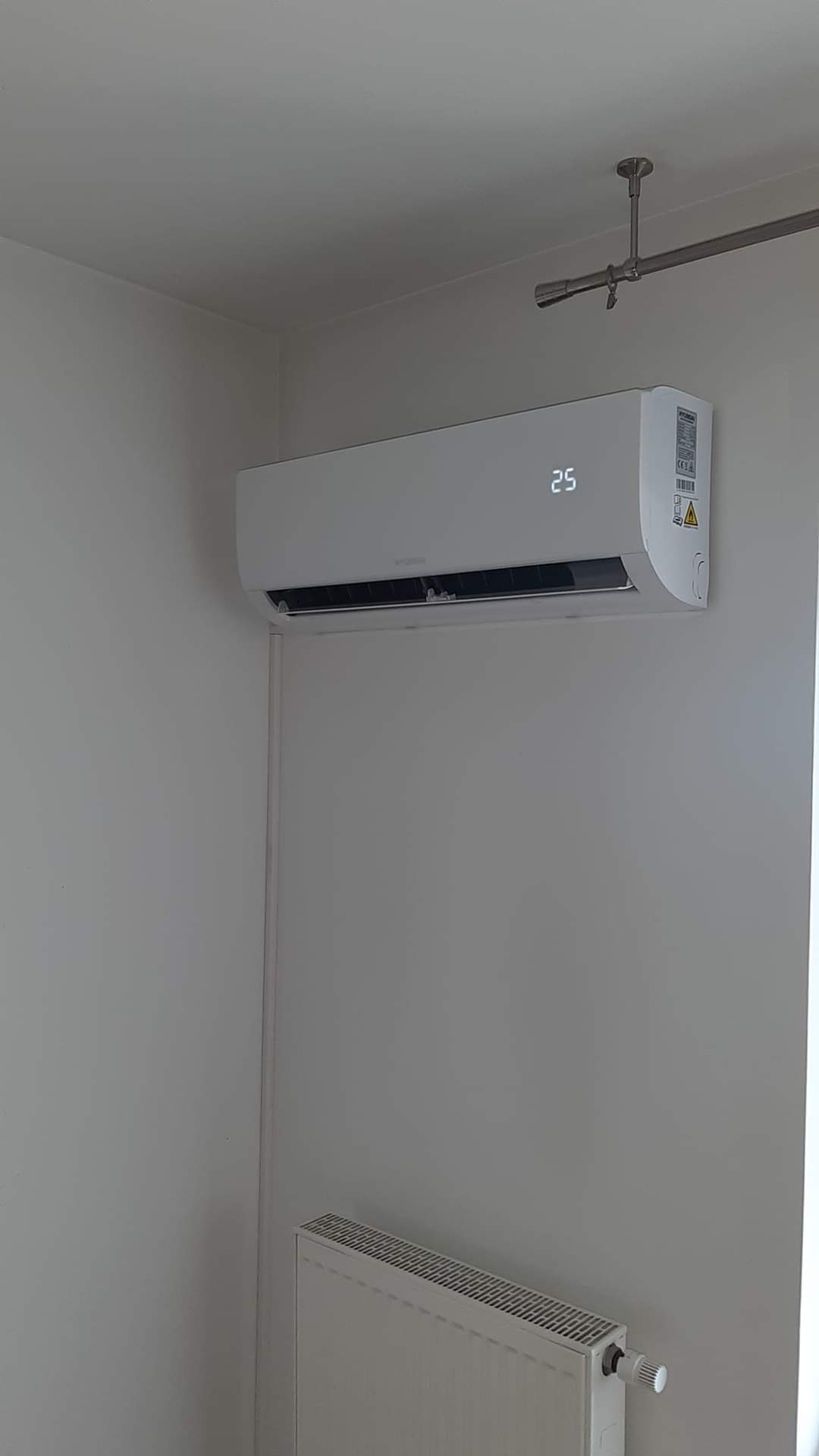 Biała klimatyzacja ścienna z wyświetlaczem temperatury 25 stopni Celsjusza, zamontowana w pomieszczeniu z grzejnikiem i szynoprzewodem oświetleniowym na suficie.