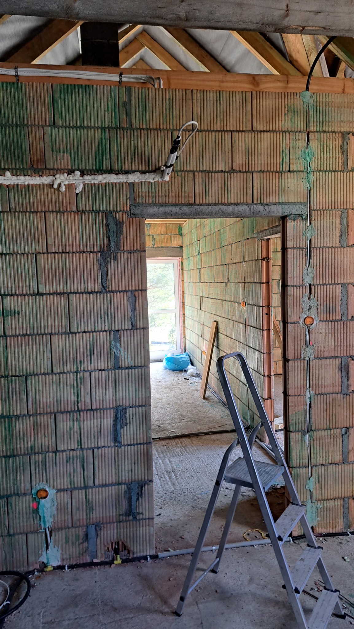 Wnętrze w trakcie budowy z widoczną ścianą z pustaków ceramicznych, częściowo otynkowaną, z poprowadzonymi przewodami elektrycznymi i rurami, drabina aluminiowa w pomieszczeniu.