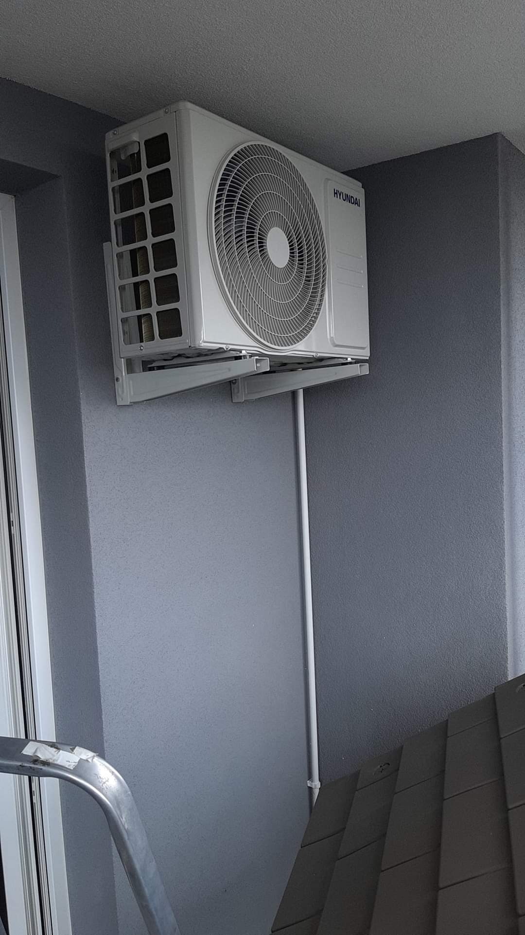 Biała jednostka klimatyzacji Hyundai zamontowana na szarej ścianie, widoczny fragment drabiny i rury instalacyjne.