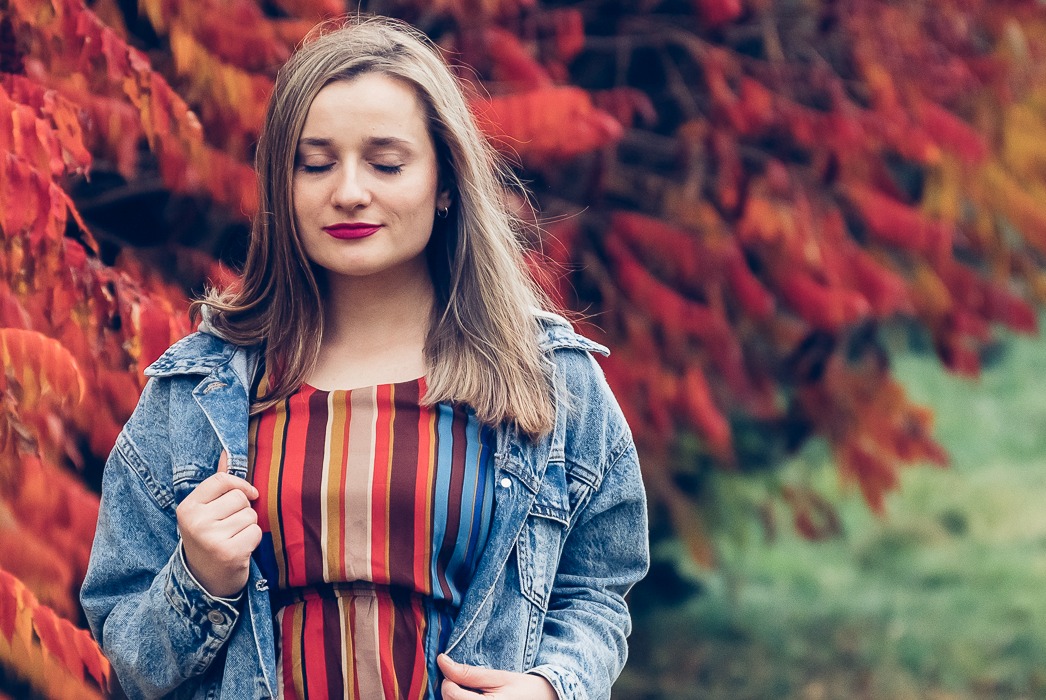 Portret kobiety z zamkniętymi oczami w dżinsowej kurtce i sukience w paski na tle jesiennych liści.