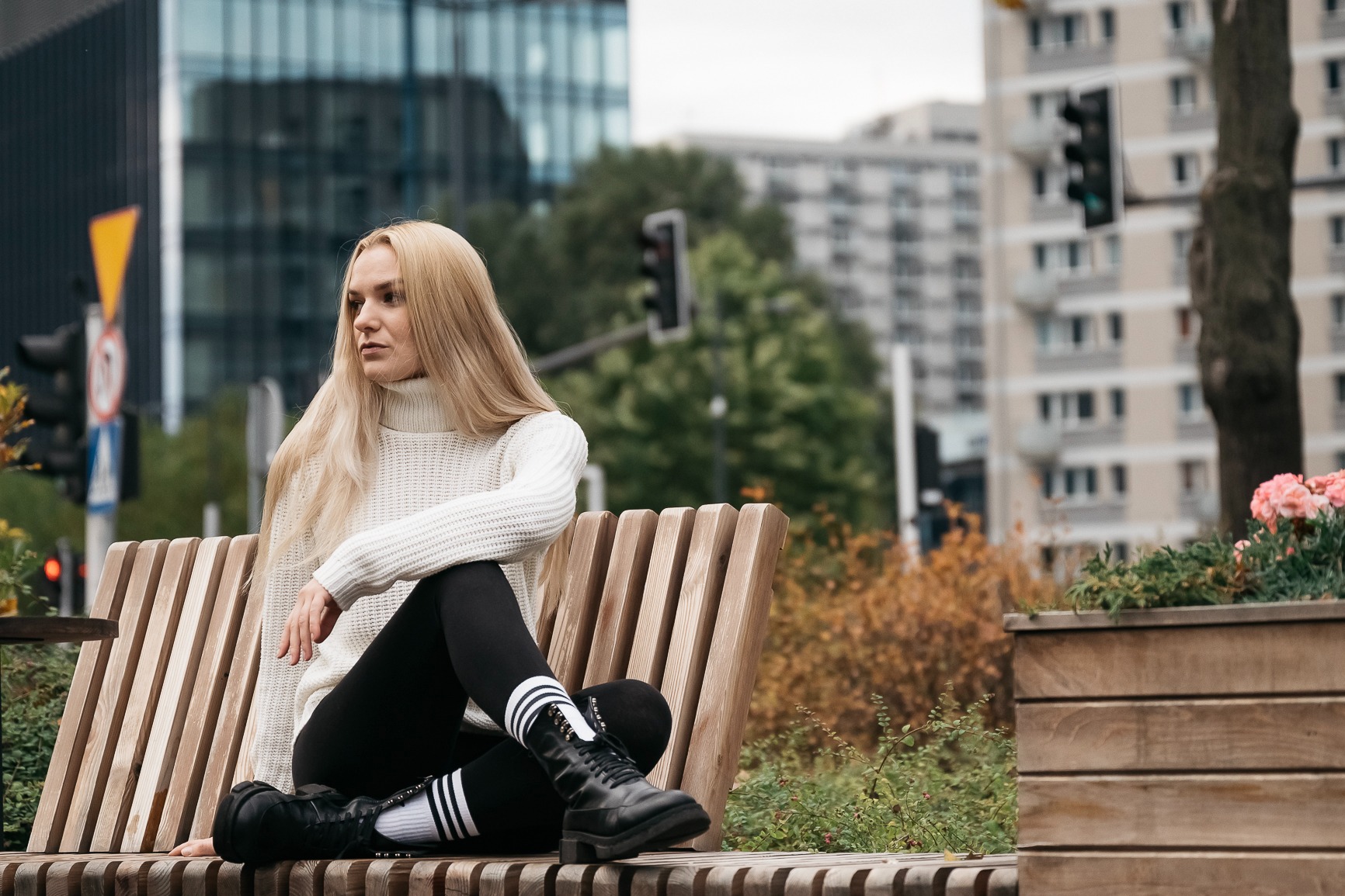 Kobieta z długimi blond włosami w białym swetrze i czarnych legginsach siedzi na drewnianej ławce w miejskim otoczeniu, z budynkami w tle i kwiatami w donicy obok.