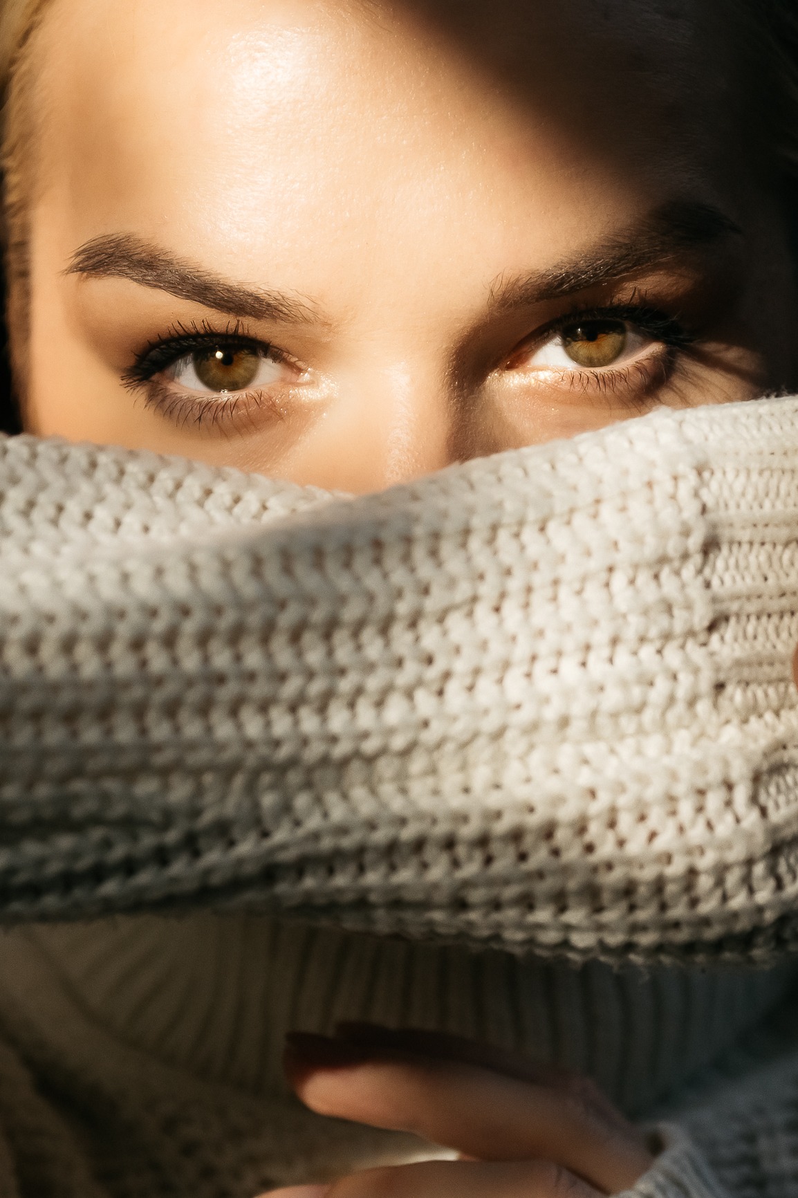 Portret kobiety z zielonymi oczami, zakrywającej twarz kremowym, grubym swetrem, oświetlona ciepłym światłem słonecznym
