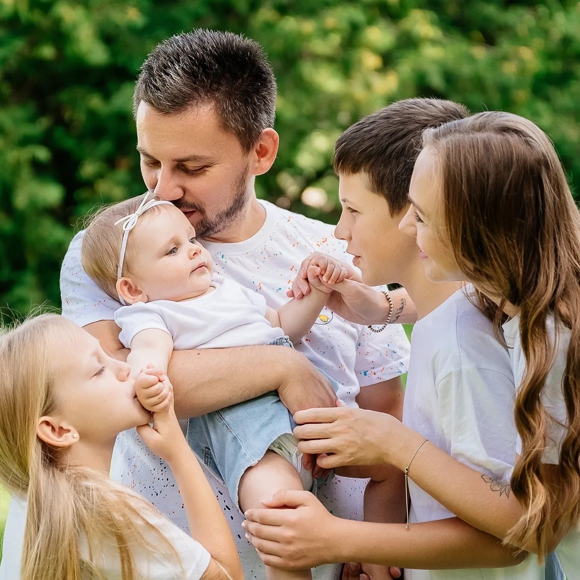Szczęśliwa rodzina: ojciec trzyma na rękach niemowlę z białą opaską, całowane w dłoń przez młodszą córkę, obok matka i syn trzymający je za rękę, wszyscy ubrani w białe koszulki na tle zieleni.