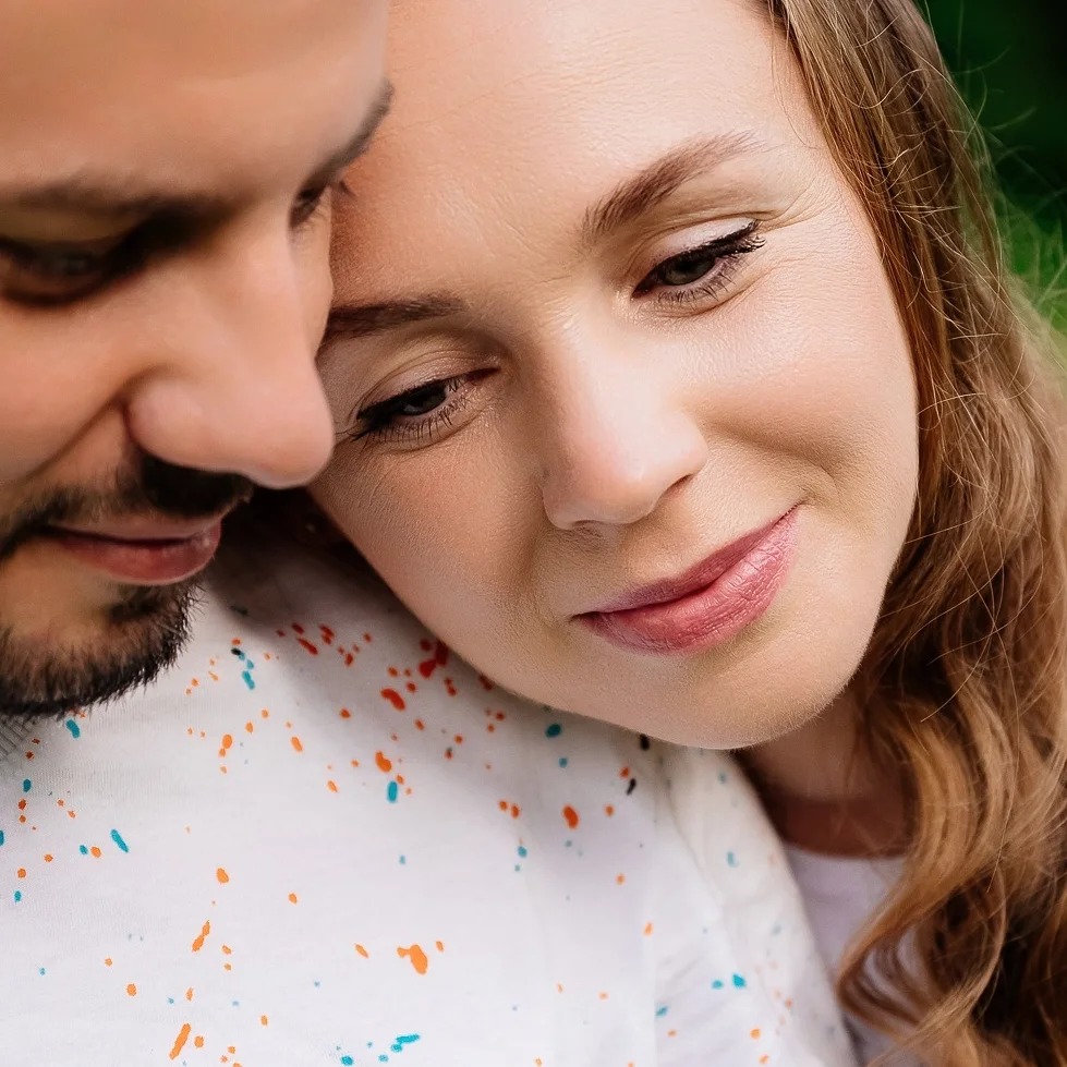 Zbliżenie na uśmiechniętą kobietę z długimi włosami, opartą głową o ramię mężczyzny z brodą, oboje ubrani w białe koszulki z kolorowymi plamkami.