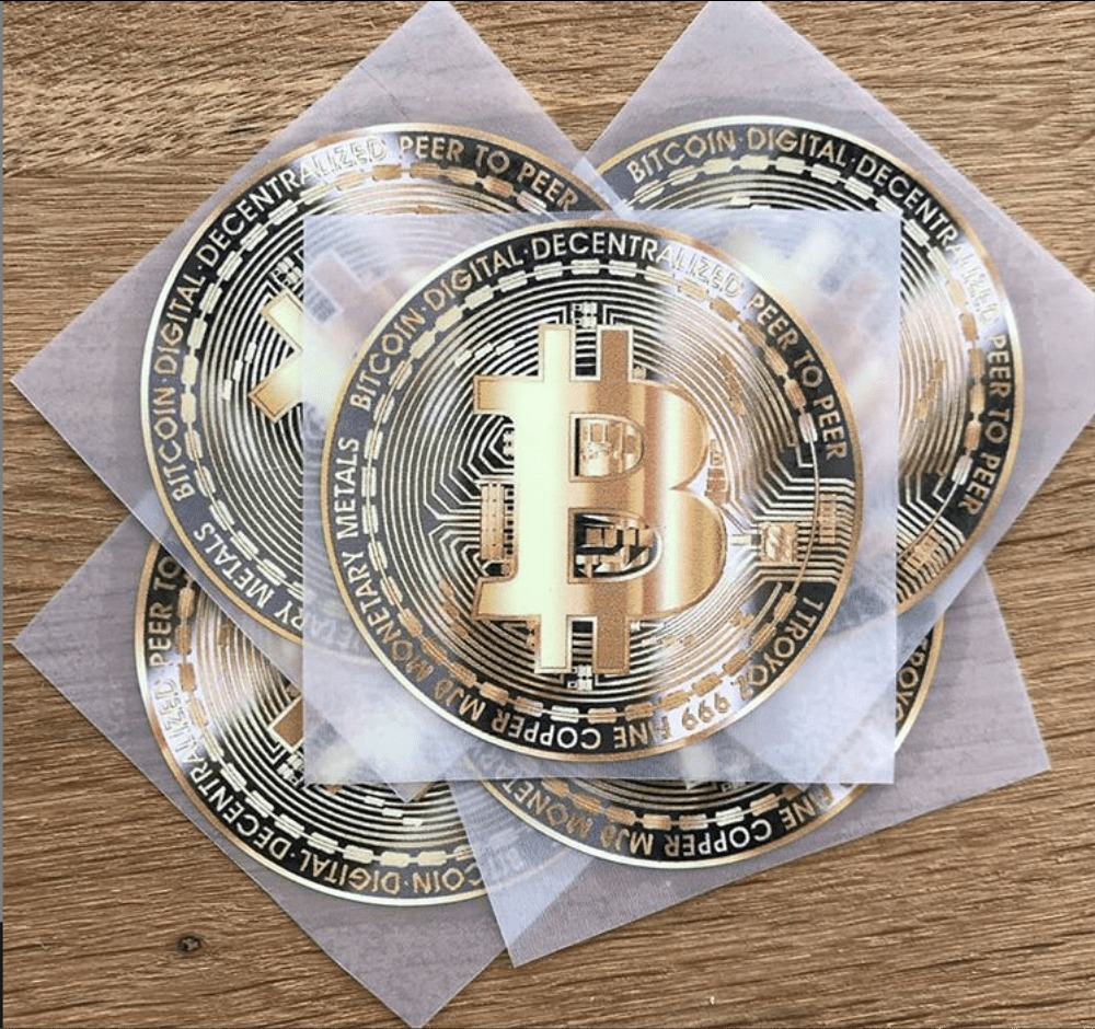 Zestaw złotych naprasowanek z motywem Bitcoin na przezroczystej folii, ułożonych na drewnianym tle.